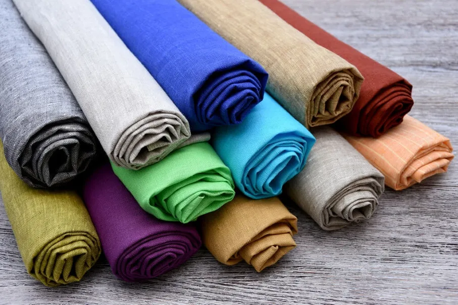 Colorful rolls of linen fabric in various shades stacked together on a wooden surface.