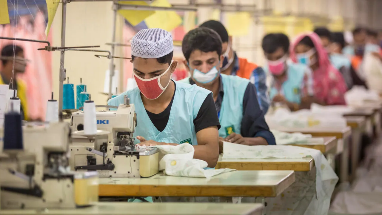 Factory workers wearing masks sewing garments on industrial machines in a clothing production line, focused on the textile manufacturing process.
