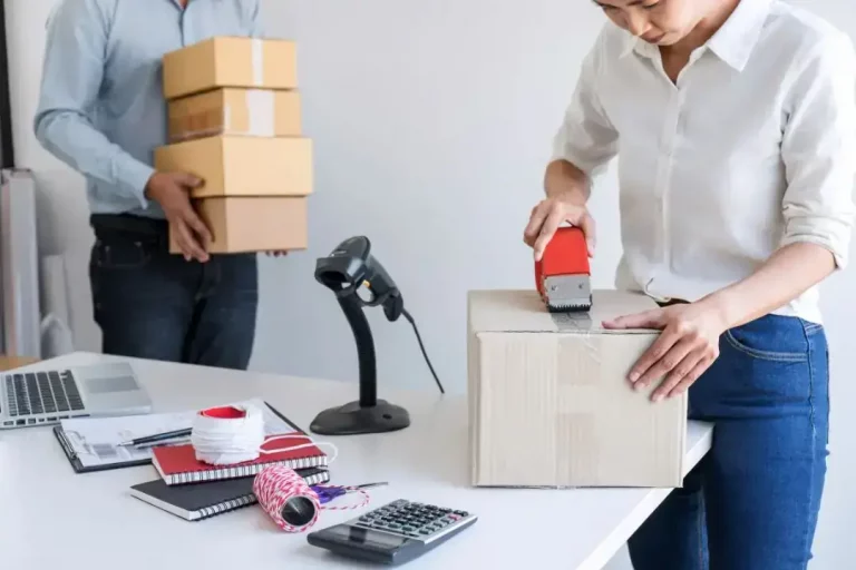 Two workers packing and sealing cardboard shipping boxes on a desk with office supplies and calculators, showing ecommerce order fulfillment and logistics.