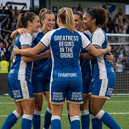Female football team huddle in jerseys with 'Greatness Begins In The Grind' quote, showing apparel branding and unity.