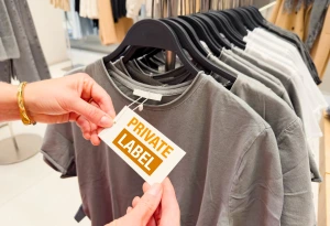 Hands attaching a “Private Label” tag to a grey t-shirt on a clothing rack in a retail store, showing branding and apparel customization.