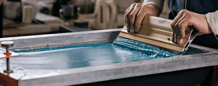 Worker using squeegee in screen printing process applying ink on mesh frame for textile or graphic design